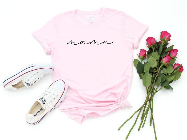 Puede incluir: Una camiseta rosa claro con la palabra "mama" en escritura negra. La imagen tambi&eacute;n presenta zapatillas blancas y un ramo de rosas rosas. La camiseta es de estilo casual.