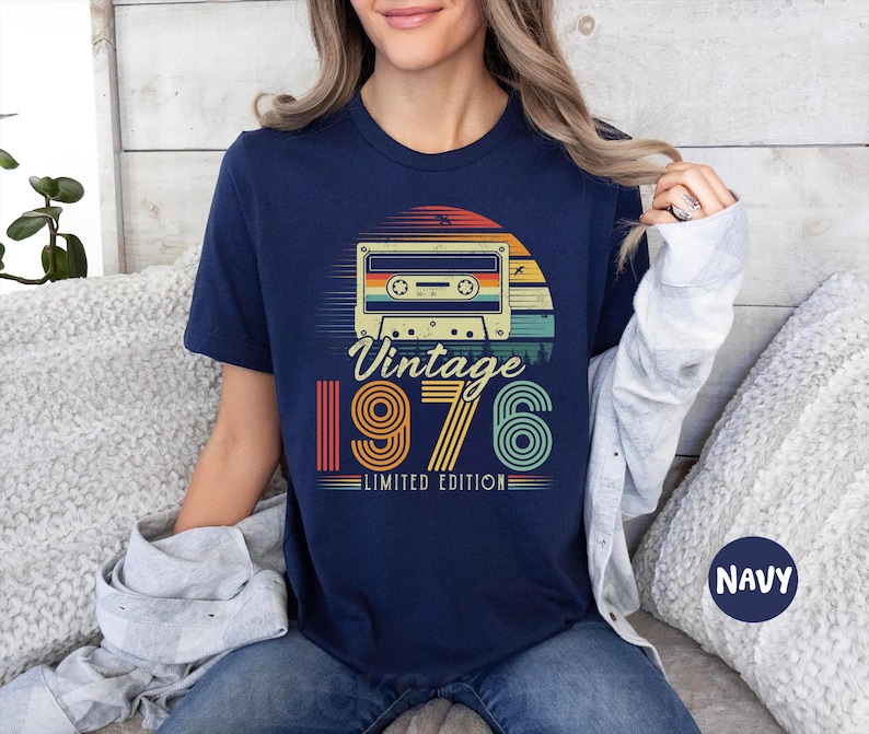 May include: A navy blue short-sleeve t-shirt featuring a graphic of a cassette tape centred above the text "Vintage 1976 LIMITED EDITION". The design incorporates a sunset motif with horizontal stripes in shades of orange, yellow, and teal.