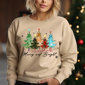 Pu&ograve; includere: Una felpa beige con un design di albero di Natale e il testo "Merry and Bright". Gli alberi sono decorati con luci colorate e stelle.