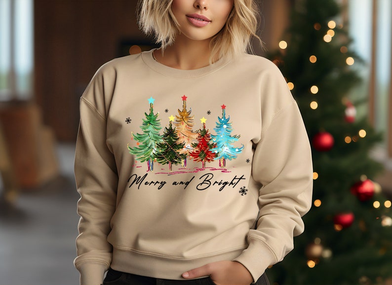 Pu&ograve; includere: Una felpa beige con un design di albero di Natale e il testo "Merry and Bright". Gli alberi sono decorati con luci colorate e stelle.