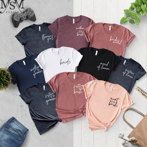 K&ouml;nnte beinhalten: Eine Auswahl an T-Shirts in verschiedenen Farben, jeweils mit einem anderen Schriftzug versehen. Die Texte lauten "Flower Girl", "Mother of the Bride", "Bridesmaid", "Bride", "Maid of Honor", "Matron of Honor", "Sister of the Groom", "Sister of the Bride" und "Aunt of the Bride".
