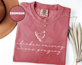May include: Crimson-colored t-shirt with a white chicken graphic and the words "chicken raising Jesus praying." The shirt has a crew neck and short sleeves. A silver necklace with a pendant is draped on the shirt.