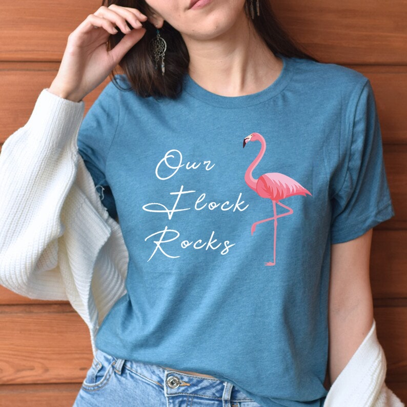 K&ouml;nnte beinhalten: T&uuml;rkisfarbenes T-Shirt mit einem rosa Flamingo-Motiv und dem Schriftzug "Our Flock Rocks" in wei&szlig;er Schrift. Der Flamingo steht auf einem Bein. Das Shirt hat kurze &Auml;rmel und einen Rundhalsausschnitt.