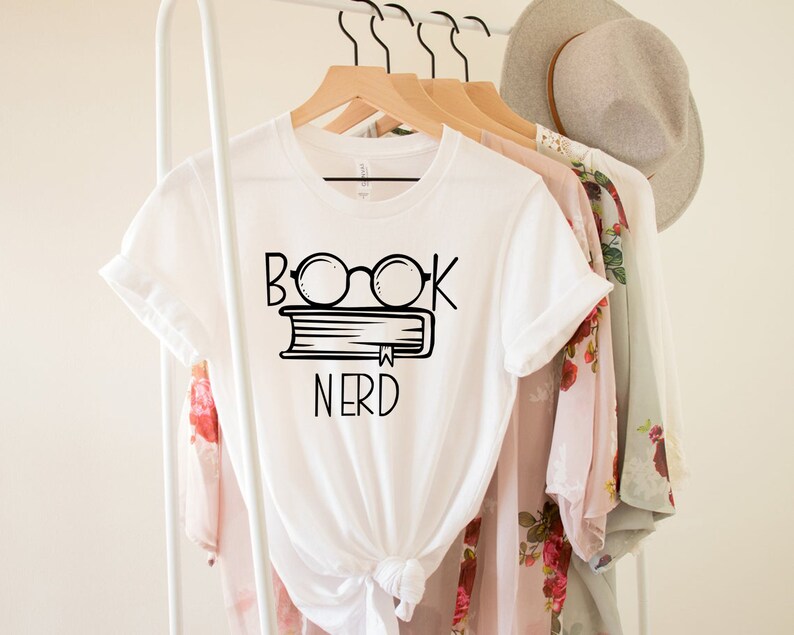 May include: White t-shirt with the words "BOOK NERD" in black, featuring glasses over a book. The shirt is hanging on a wooden hanger with other clothing items. The design is simple and bold, perfect for book lovers.