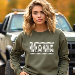 Puede incluir: Una sudadera verde oliva con la palabra "MAMA" en un dise&ntilde;o a cuadros blancos. La sudadera es de cuello redondo y la lleva puesta una persona. El fondo incluye un veh&iacute;culo verde y follaje oto&ntilde;al.