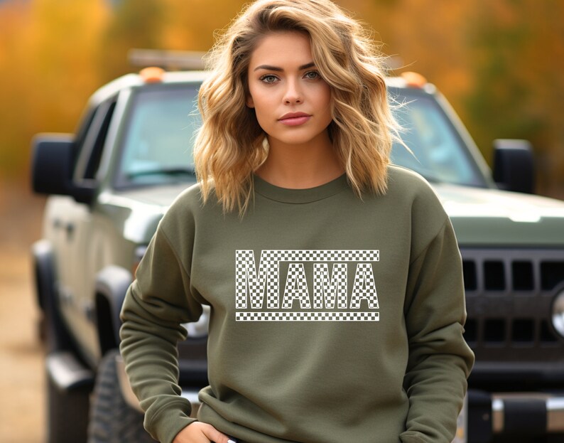 Puede incluir: Una sudadera verde oliva con la palabra "MAMA" en un dise&ntilde;o a cuadros blancos. La sudadera es de cuello redondo y la lleva puesta una persona. El fondo incluye un veh&iacute;culo verde y follaje oto&ntilde;al.