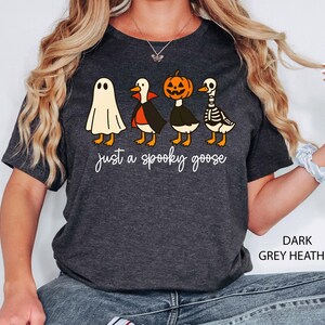 Puede incluir: Camiseta gris oscuro jaspeado con un dise&ntilde;o tem&aacute;tico de Halloween. La camiseta muestra cuatro gansos de dibujos animados disfrazados de fantasma, vampiro, calabaza y esqueleto. El texto "just a spooky goose" est&aacute; escrito debajo.