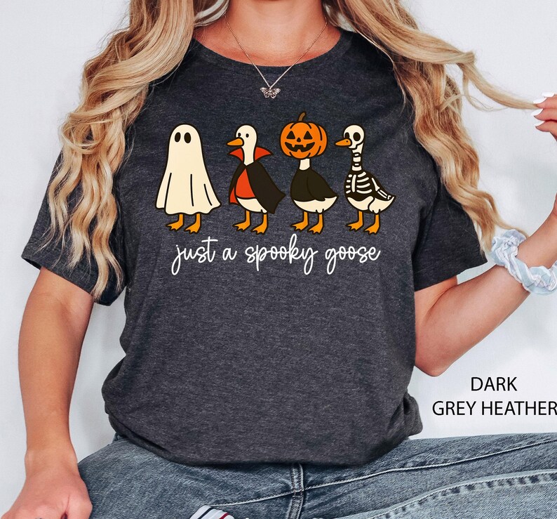 Puede incluir: Camiseta gris oscuro jaspeado con un dise&ntilde;o tem&aacute;tico de Halloween. La camiseta muestra cuatro gansos de dibujos animados disfrazados de fantasma, vampiro, calabaza y esqueleto. El texto "just a spooky goose" est&aacute; escrito debajo.