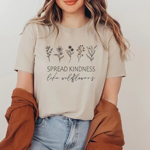Op de afbeelding: Een beige T-shirt met een rij eenvoudige zwarte wilde bloemen en de tekst "SPREAD KINDNESS like wildflowers".