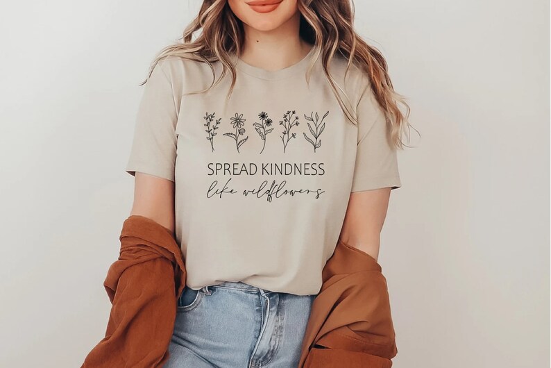 Op de afbeelding: Een beige T-shirt met een rij eenvoudige zwarte wilde bloemen en de tekst "SPREAD KINDNESS like wildflowers".