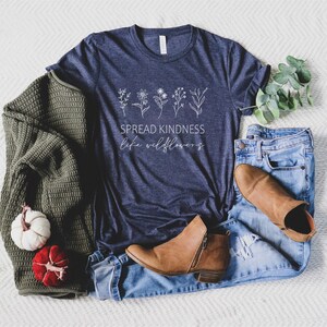 Op de afbeelding: Een marineblauw t-shirt met een witte afbeelding van wilde bloemen en de tekst "Spread Kindness Like Wildflowers". Het shirt ligt op een wit oppervlak met een groene trui, jeans en bruine laarzen.
