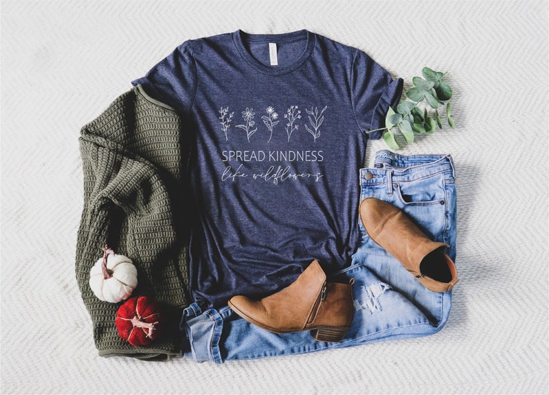 Op de afbeelding: Een marineblauw t-shirt met een witte afbeelding van wilde bloemen en de tekst "Spread Kindness Like Wildflowers". Het shirt ligt op een wit oppervlak met een groene trui, jeans en bruine laarzen.