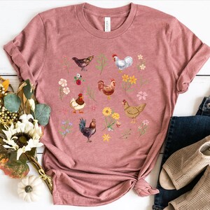 May include: A pink t-shirt with a graphic print of chickens and flowers. The chickens are in various colors, including white, brown, and black. The flowers are in shades of pink, yellow, and purple.