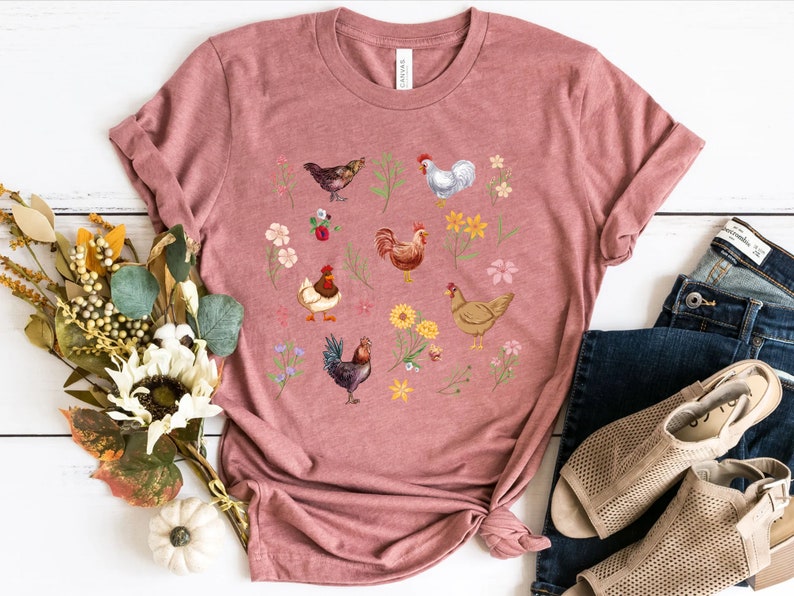 May include: A pink t-shirt with a graphic print of chickens and flowers. The chickens are in various colors, including white, brown, and black. The flowers are in shades of pink, yellow, and purple.