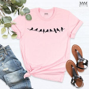 May include: A pink t-shirt with a graphic of a line of black birds on a wire. The shirt is made of a soft, comfortable material and has a crew neck.