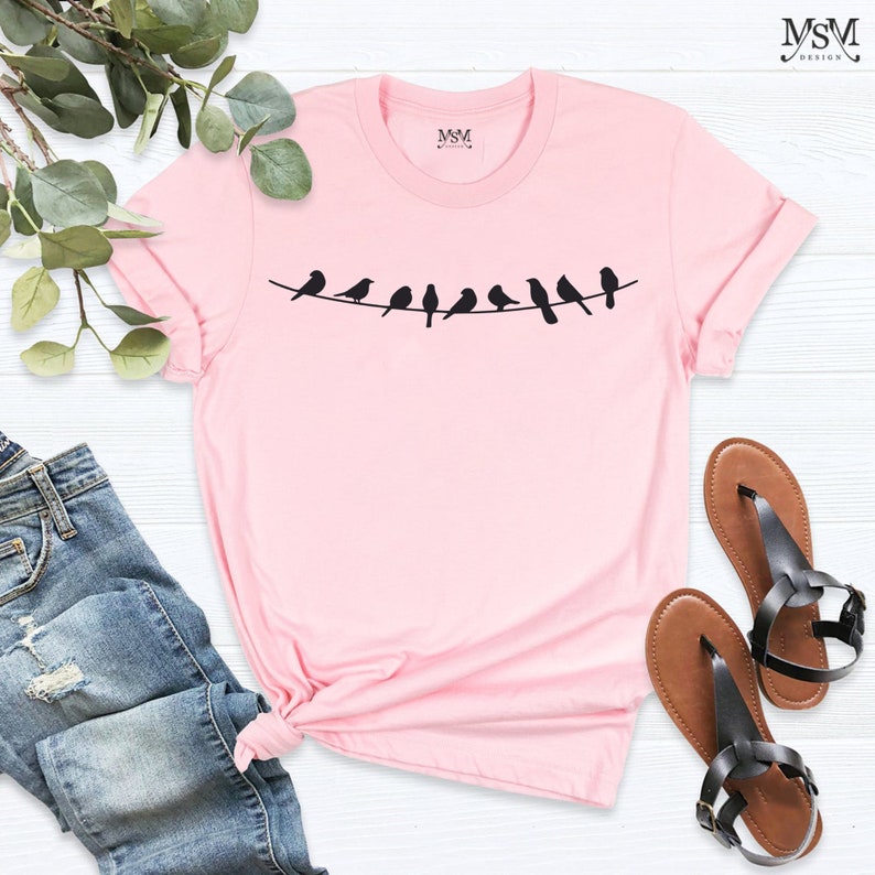 May include: A pink t-shirt with a graphic of a line of black birds on a wire. The shirt is made of a soft, comfortable material and has a crew neck.