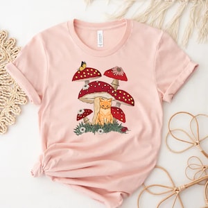 K&ouml;nnte beinhalten: Hellrosa T-Shirt mit einer verspielten Illustration. Das Design zeigt eine rot getigerte Katze unter roten Pilzen mit wei&szlig;en Punkten, einen Schmetterling und kleine Blumen. Das T-Shirt ist aus weichem Material.