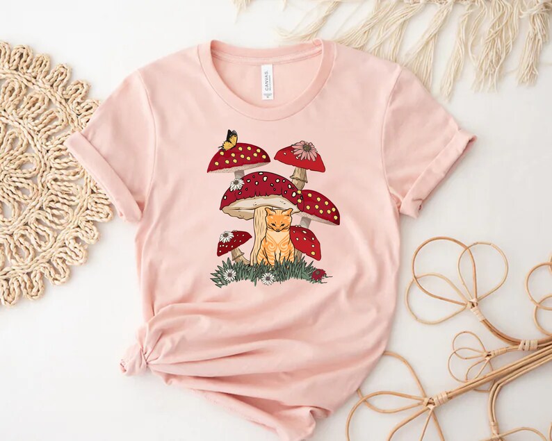 K&ouml;nnte beinhalten: Hellrosa T-Shirt mit einer verspielten Illustration. Das Design zeigt eine rot getigerte Katze unter roten Pilzen mit wei&szlig;en Punkten, einen Schmetterling und kleine Blumen. Das T-Shirt ist aus weichem Material.