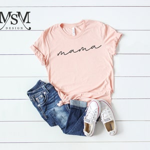 Puede incluir: Una camiseta rosa claro con la palabra "mama" en escritura negra. La camiseta se combina con vaqueros azules y zapatillas de lona blancas. Los art&iacute;culos est&aacute;n colocados sobre una superficie de madera blanca.