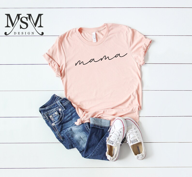 Puede incluir: Una camiseta rosa claro con la palabra "mama" en escritura negra. La camiseta se combina con vaqueros azules y zapatillas de lona blancas. Los art&iacute;culos est&aacute;n colocados sobre una superficie de madera blanca.