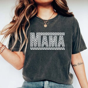Puede incluir: Camiseta gris oscuro con la palabra "MAMA" en letras blancas a cuadros. La palabra est&aacute; centrada y enmarcada por un borde a cuadros. La camiseta tiene un aspecto vintage y desgastado.