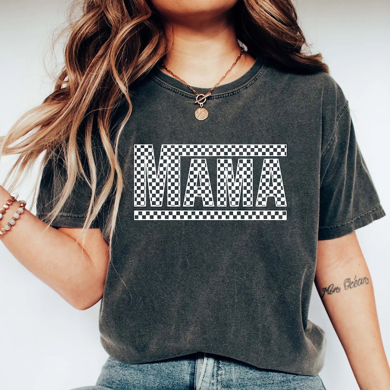 Puede incluir: Camiseta gris oscuro con la palabra "MAMA" en letras blancas a cuadros. La palabra est&aacute; centrada y enmarcada por un borde a cuadros. La camiseta tiene un aspecto vintage y desgastado.