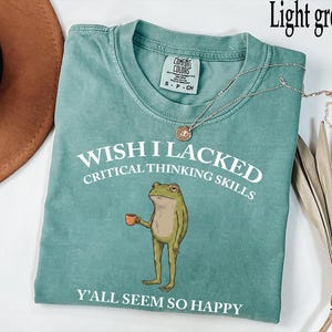 May include: A light green short-sleeve t-shirt featuring a graphic of a standing frog holding a small orange mug. The white text reads WISH I LACKED CRITICAL THINKING SKILLS and Y'ALL SEEM SO HAPPY. The shirt has a crew neckline and a brand label.