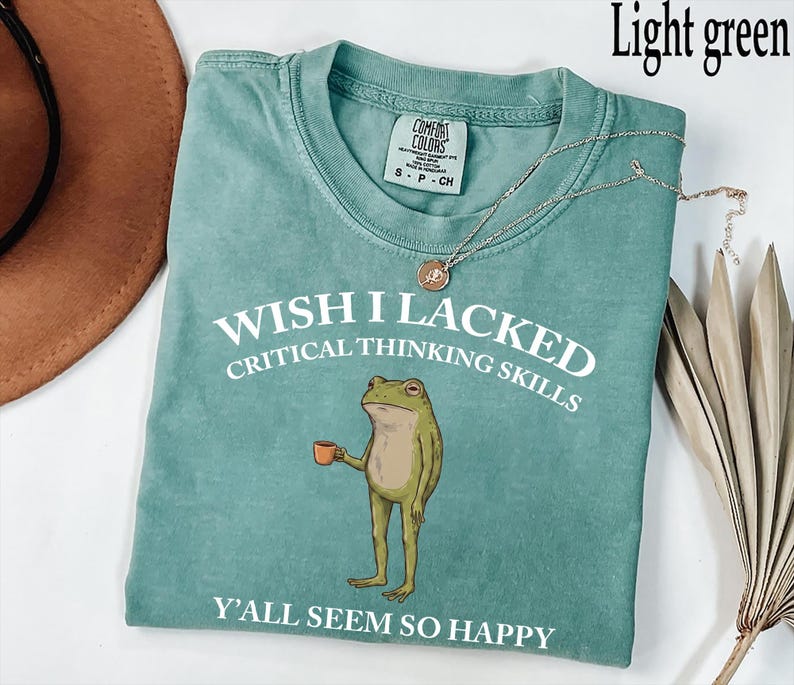 May include: A light green short-sleeve t-shirt featuring a graphic of a standing frog holding a small orange mug. The white text reads WISH I LACKED CRITICAL THINKING SKILLS and Y'ALL SEEM SO HAPPY. The shirt has a crew neckline and a brand label.