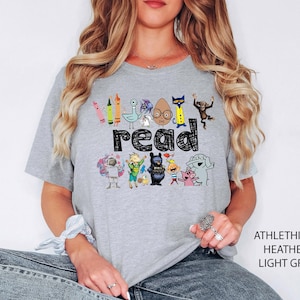 May include: A light grey heather t-shirt featuring the word read in a black, hand-drawn style font. The design includes colorful illustrations of various children's book characters, including a pigeon, a cat, a monkey, and an elephant.