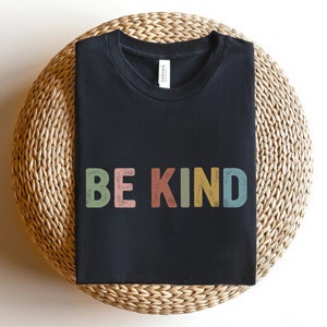 May include: A black t-shirt with the words "BE KIND" printed across the front in colorful, retro-style lettering. The letters are in shades of green, pink, yellow, and blue. The shirt is displayed on a woven, circular surface.