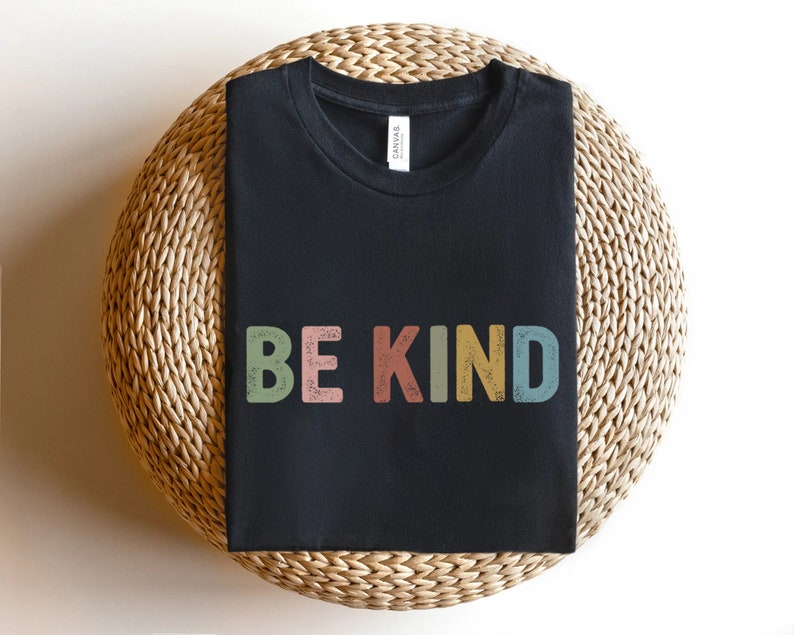 May include: A black t-shirt with the words "BE KIND" printed across the front in colorful, retro-style lettering. The letters are in shades of green, pink, yellow, and blue. The shirt is displayed on a woven, circular surface.