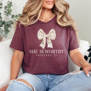 May include: Burgundy t-shirt with a cream-colored bow graphic and the text "SHE IS WORTHY PROVERBS 31" in white. The shirt has short sleeves and a crew neck.