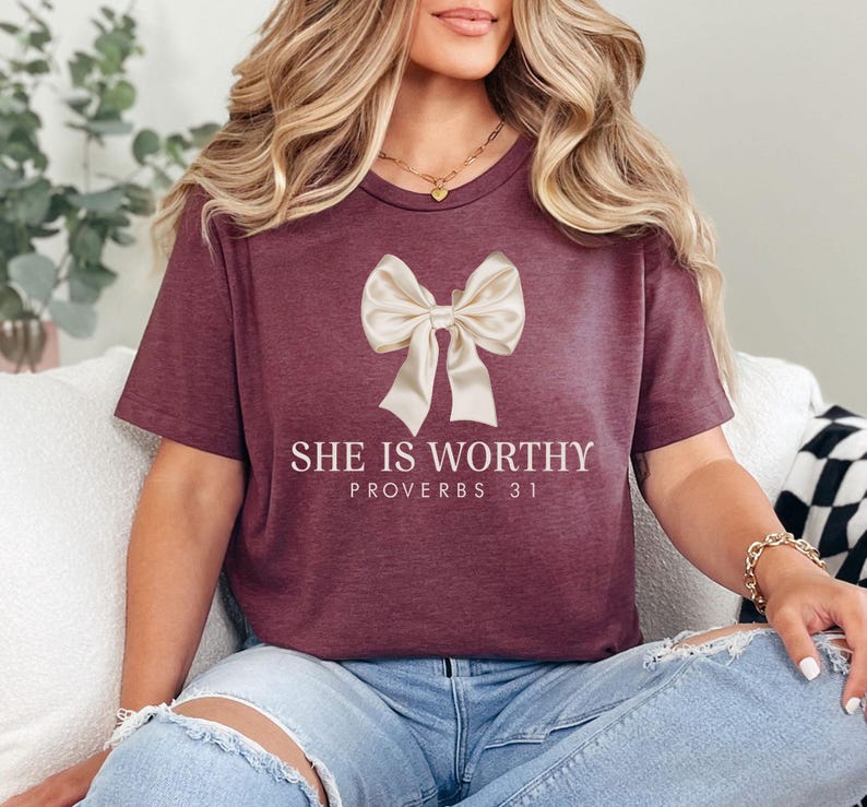 May include: Burgundy t-shirt with a cream-colored bow graphic and the text "SHE IS WORTHY PROVERBS 31" in white. The shirt has short sleeves and a crew neck.