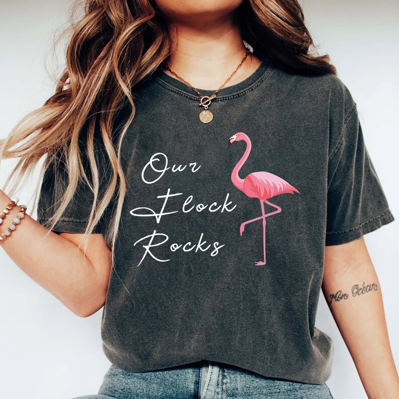 K&ouml;nnte beinhalten: Dunkelgraues T-Shirt mit einem rosa Flamingo-Motiv und dem wei&szlig;en Text "Our Flock Rocks". Das Shirt hat einen Vintage-Look und ist aus einem weichen, bequemen Material gefertigt. Ein l&auml;ssiges T-Shirt f&uuml;r den Alltag.