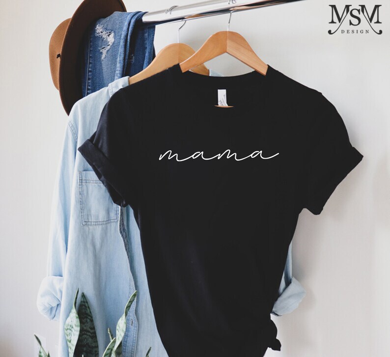 Puede incluir: Camiseta negra con la palabra "mama" en blanco en fuente cursiva. La camiseta est&aacute; colgada en una percha de madera, con una camisa vaquera azul claro y un sombrero marr&oacute;n en el fondo.