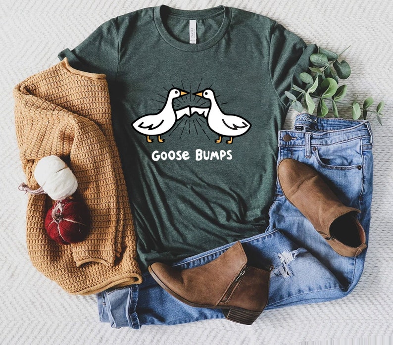 May include: A green t-shirt with a white goose graphic and the text "Goose Bumps". The shirt is displayed on a white surface with a brown sweater, blue jeans, and brown boots.