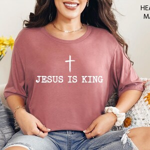 K&ouml;nnte beinhalten: Heather-Mauve-T-Shirt mit dem wei&szlig;en Text "JESUS IS KING" und einem wei&szlig;en Kreuz dar&uuml;ber. Das Shirt ist einfarbig und hat kurze &Auml;rmel. Das Model tr&auml;gt das Shirt.