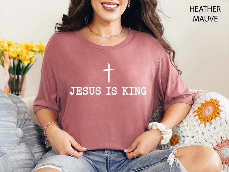 K&ouml;nnte beinhalten: Heather-Mauve-T-Shirt mit dem wei&szlig;en Text "JESUS IS KING" und einem wei&szlig;en Kreuz dar&uuml;ber. Das Shirt ist einfarbig und hat kurze &Auml;rmel. Das Model tr&auml;gt das Shirt.