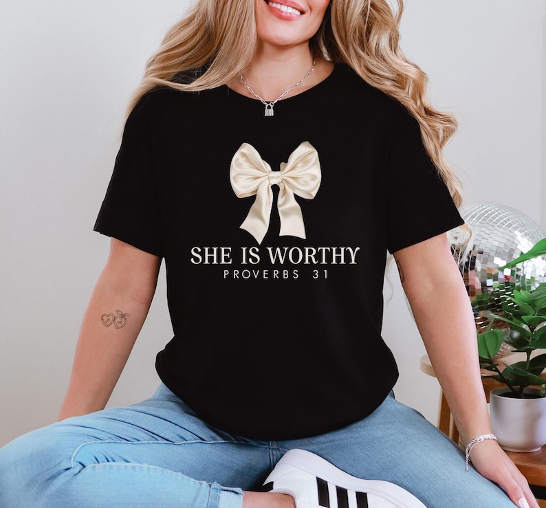 May include: Black t-shirt featuring a cream-colored bow graphic and the text "SHE IS WORTHY PROVERBS 31" in white. The shirt has a classic crew neck design.