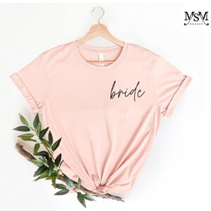 May include: A blush pink t-shirt with the word "bride" written in black script on the front.