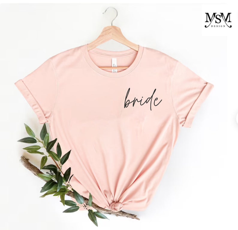 May include: A blush pink t-shirt with the word "bride" written in black script on the front.