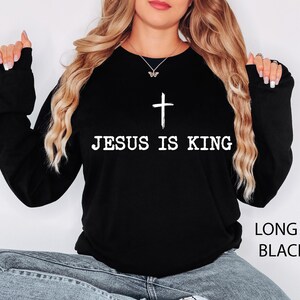 K&ouml;nnte beinhalten: Schwarzes Langarm-Sweatshirt mit dem wei&szlig;en Text &bdquo;JESUS IS KING&ldquo; und einem wei&szlig;en Kreuz. Das Kleidungsst&uuml;ck hat eine einfarbige schwarze Farbe.