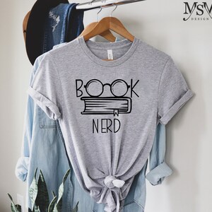 May include: A heather gray t-shirt with a black graphic of glasses over a book and the word "NERD" below. The shirt is hanging on a wooden hanger. A denim shirt and a hat are in the background.