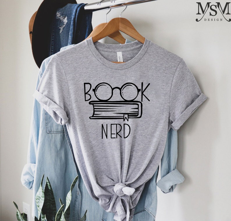 May include: A heather gray t-shirt with a black graphic of glasses over a book and the word "NERD" below. The shirt is hanging on a wooden hanger. A denim shirt and a hat are in the background.