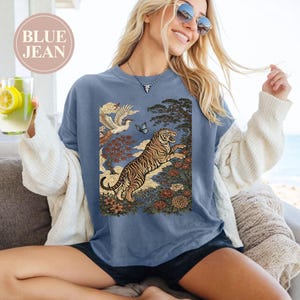 May include: A blue crewneck t-shirt featuring a graphic print of a tiger leaping among flowers, trees, and a bird in flight. The text "BLUE JEAN" is printed in a circular badge in the upper left corner. The shirt has short sleeves and a round neckline.