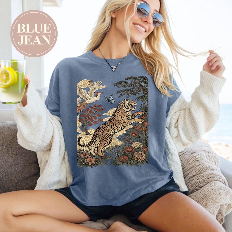 May include: A blue crewneck t-shirt featuring a graphic print of a tiger leaping among flowers, trees, and a bird in flight. The text "BLUE JEAN" is printed in a circular badge in the upper left corner. The shirt has short sleeves and a round neckline.