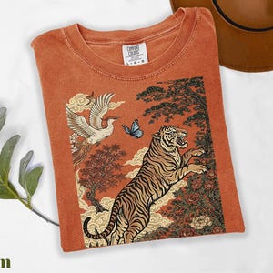 May include: A rust-colored t-shirt featuring a graphic print of a tiger, a crane in flight, a blue butterfly, and stylized trees and clouds. The shirt has a crew neckline and a Comfort Colors brand label on the inside collar indicating a size large.