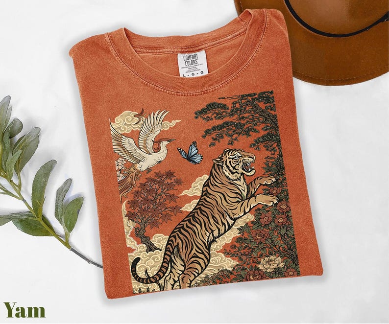 May include: A rust-colored t-shirt featuring a graphic print of a tiger, a crane in flight, a blue butterfly, and stylized trees and clouds. The shirt has a crew neckline and a Comfort Colors brand label on the inside collar indicating a size large.