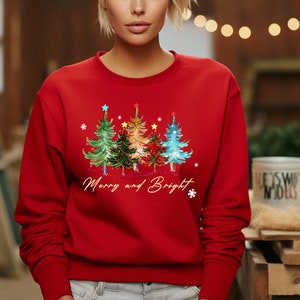 Pu&ograve; includere: Felpa rossa di Natale con un design di tre alberi di Natale colorati e il testo "Merry and Bright".