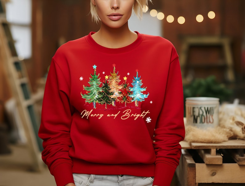 Pu&ograve; includere: Felpa rossa di Natale con un design di tre alberi di Natale colorati e il testo "Merry and Bright".